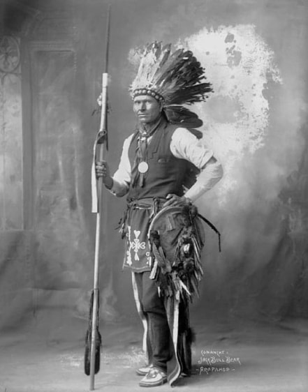 Studio portrait of Jock Bull Bear, Arapaho, holding a staff